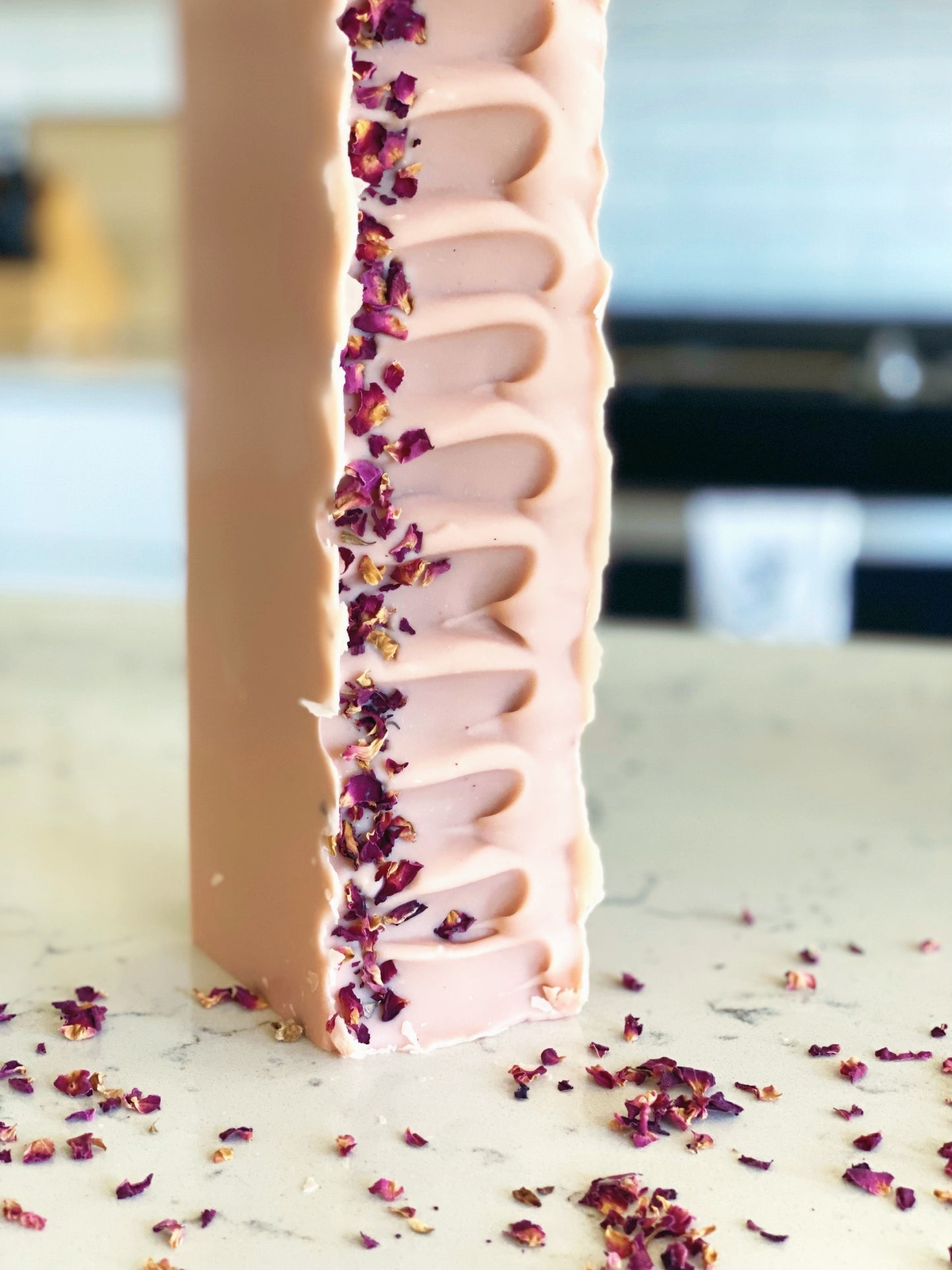 one loaf of coconut and rose soap standing vertically on a quartz countertop in a white kitchen. there are rose petals sprinkled on the counter.