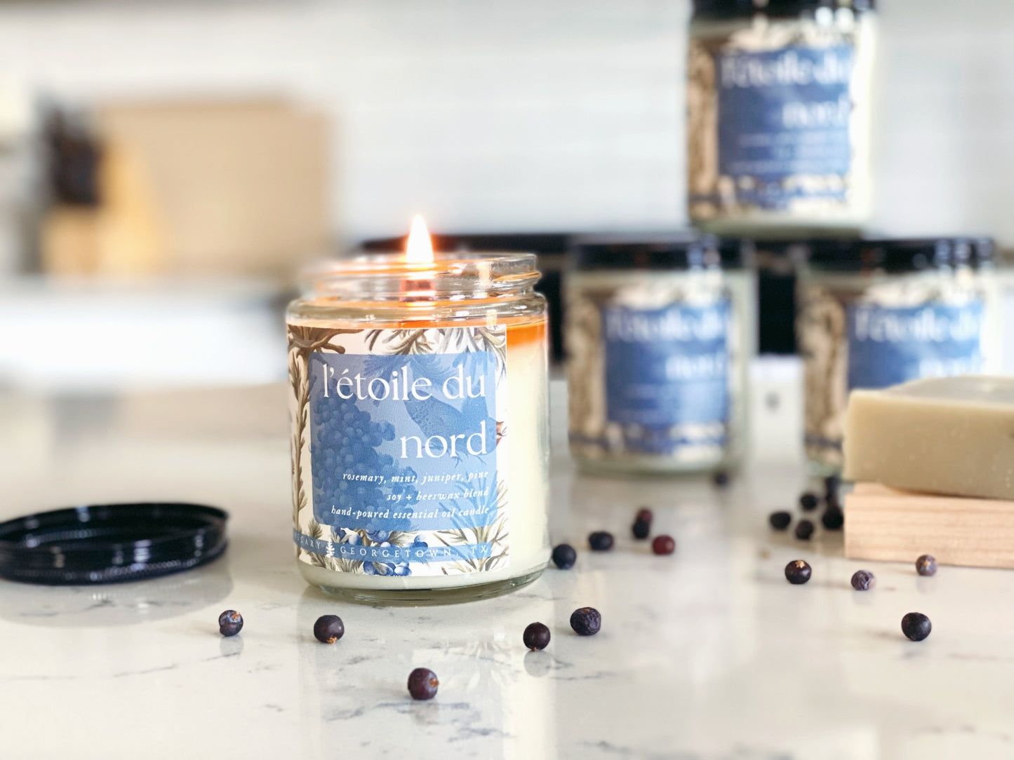 four jars of l'etoile du nord candles sitting on a quartz countertop in a white kitchen. the candle in the foreground is lit and there are dried juniper berries on the counter.