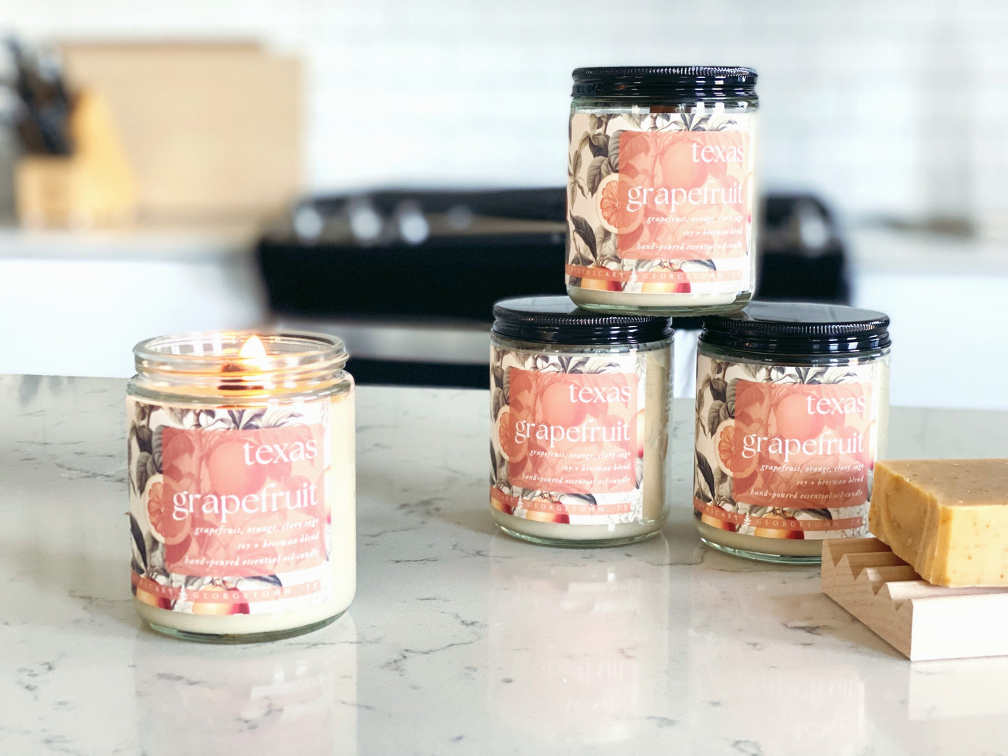 four jars of texas grapefruit candles on a quartz countertop in a white kitchen. the candle in the foreground is lit and there is a texas grapefruit bar of soap in the background.