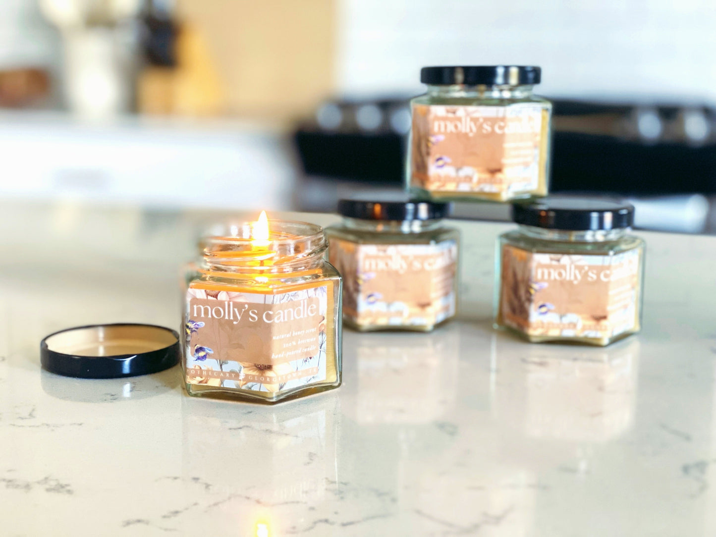 four jars of Molly's Candle 100% beeswax on a quartz countertop in a white kitchen. the candle in the foreground is lit.