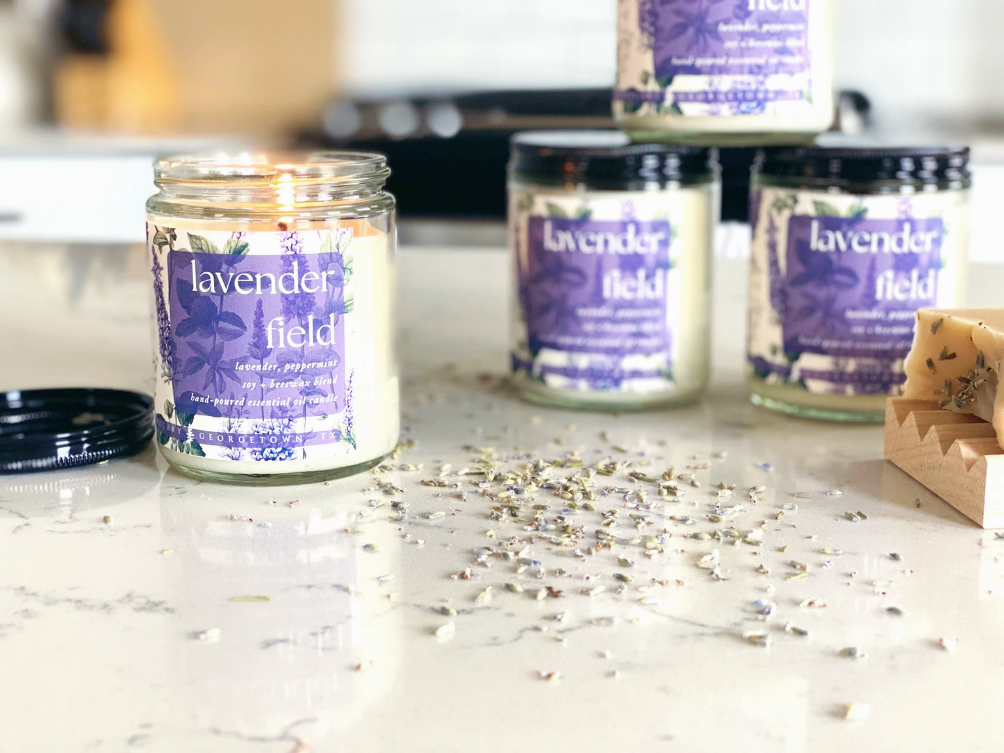four lavender field candle jars are sitting on a quartz countertop in a white kitchen. the candle in the foreground is lit and there are dried lavender buds sprinkled on the counter.