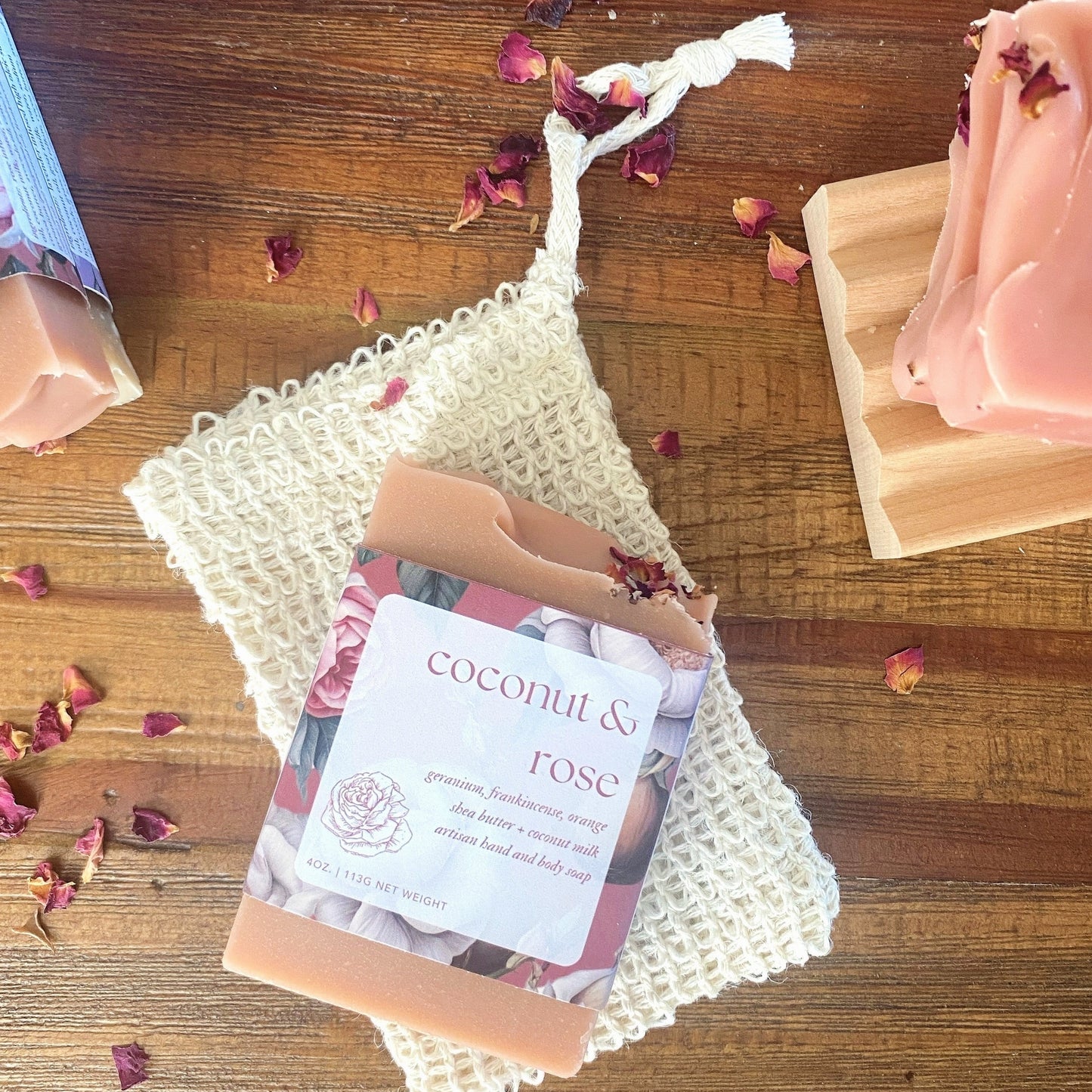 overhead photo of coconut and rose soap on a wooden table with an agave fiber soap saver and rose petals sprinkled on top.
