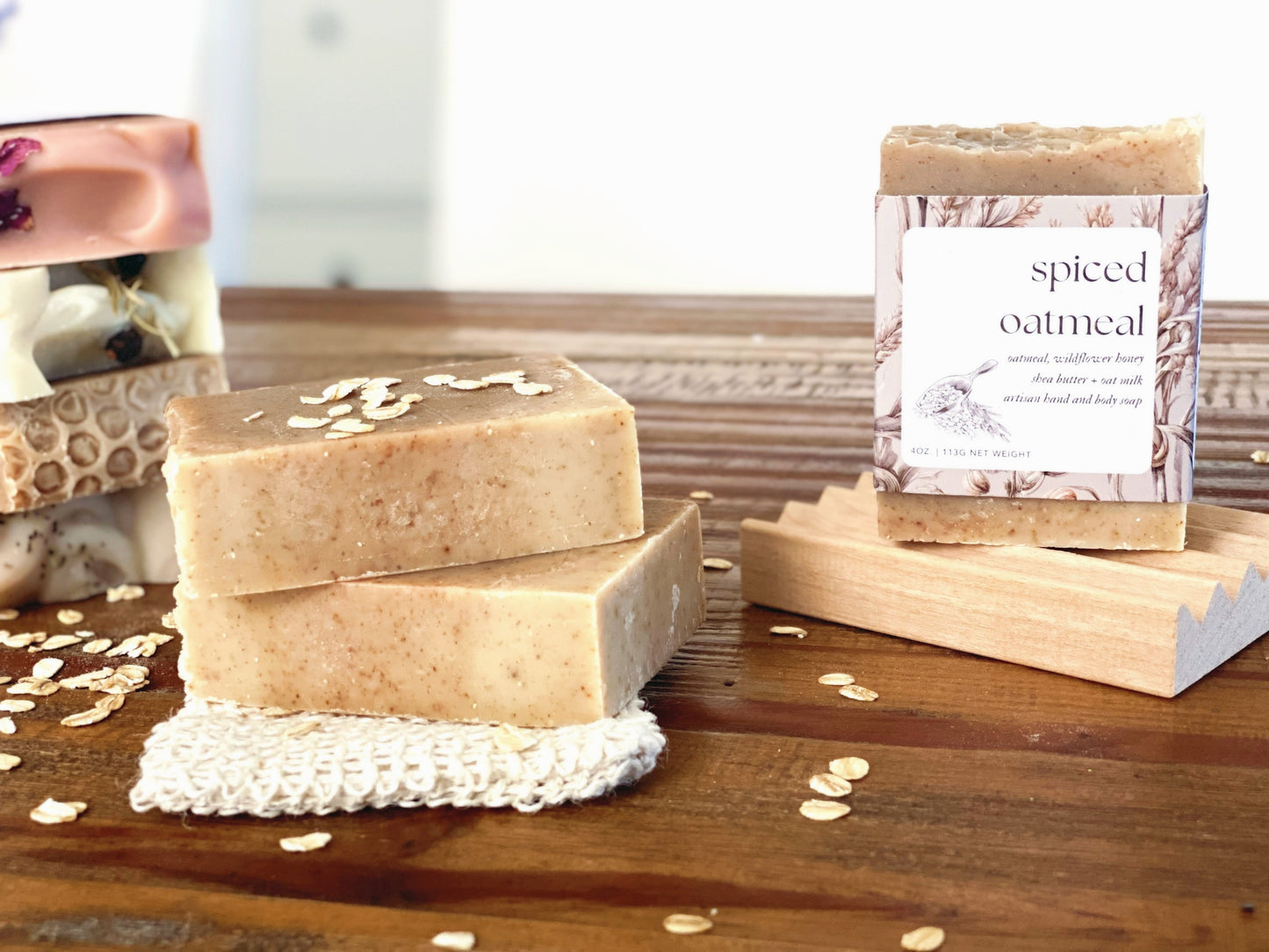 spiced oatmeal soap stacked on wooden table and wooden soap tray. there is a stack of soap assortment in the background with whole oats sprinkled on top.
