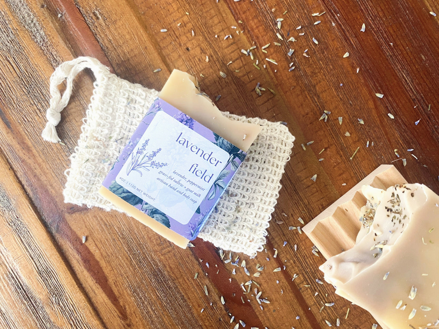 overhead photo of lavender field soap with product labeling on a sisal soap saver sitting on a wooden table. there are dried lavender buds sprinkled on top.