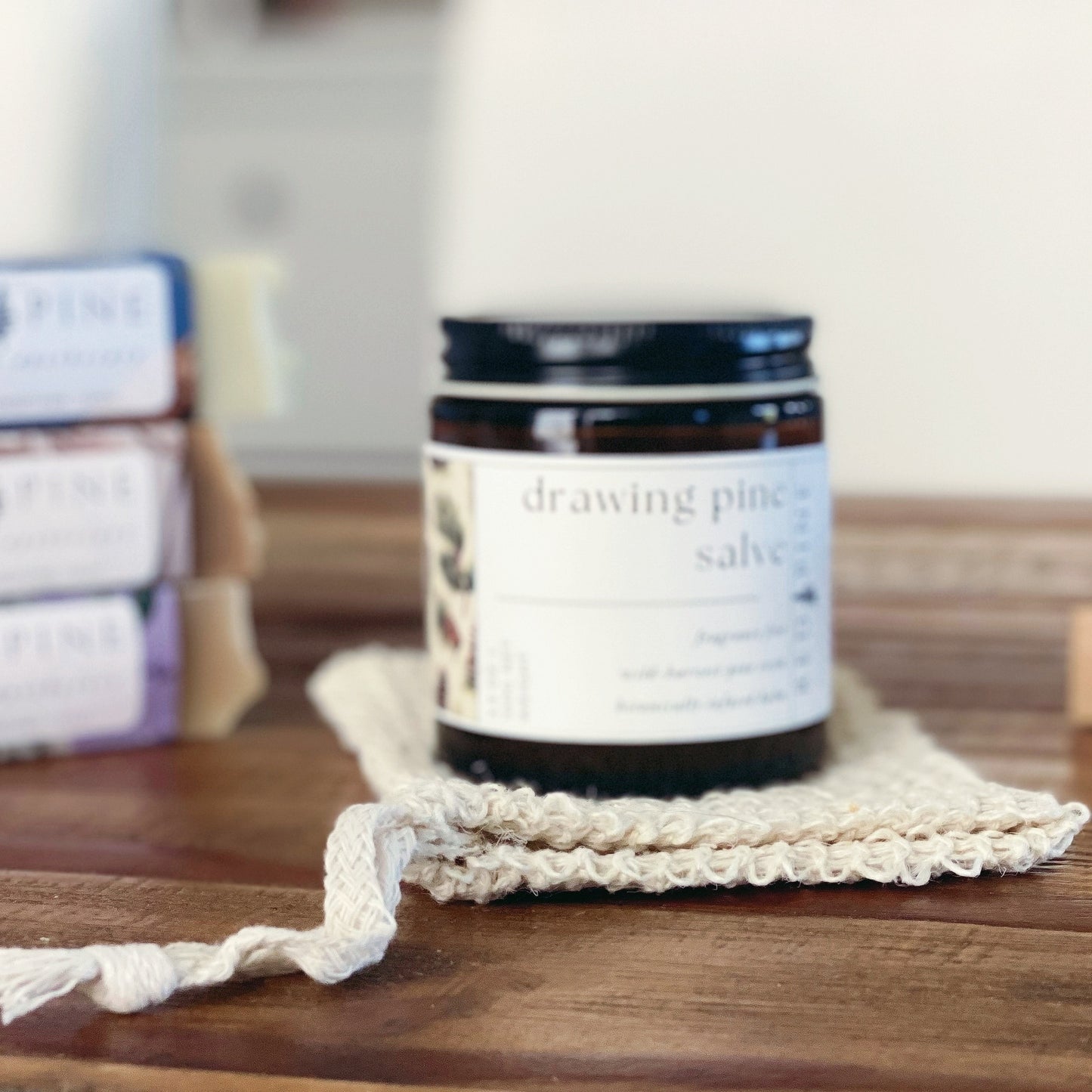 amber jar of drawing pine salve on a wooden table with a stack of soaps in the background.