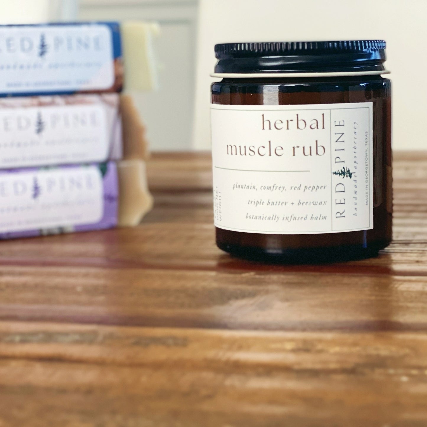 glass jar of herbal muscle rub on a wooden table. there is a stack of soaps in the background.