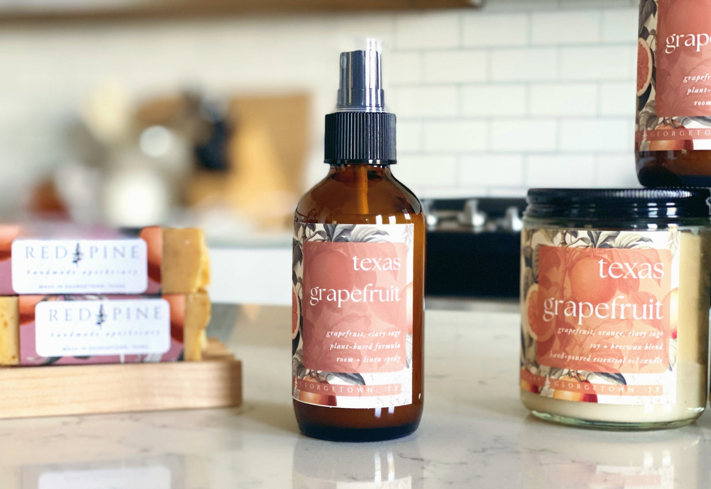 bottle of texas grapefruit room and linen spray on a quartz countertop in a white kitchen. there are texas grapefruit soaps and candles in the background.