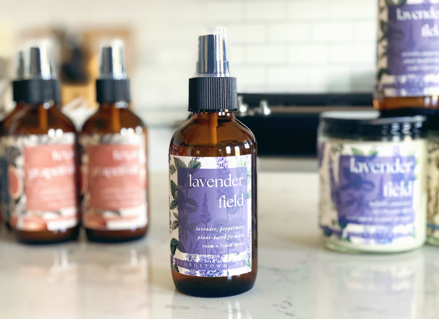 lavender field room spray bottle on quartz countertop in a white kitchen. there are lavender field candles and texas grapefruit room sprays in the background.