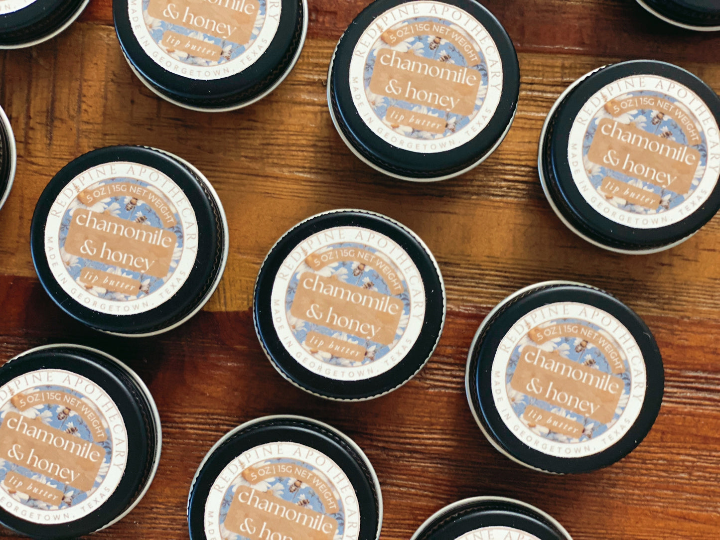 nine tins of chamomile and honey lip butter laying on a wooden table.