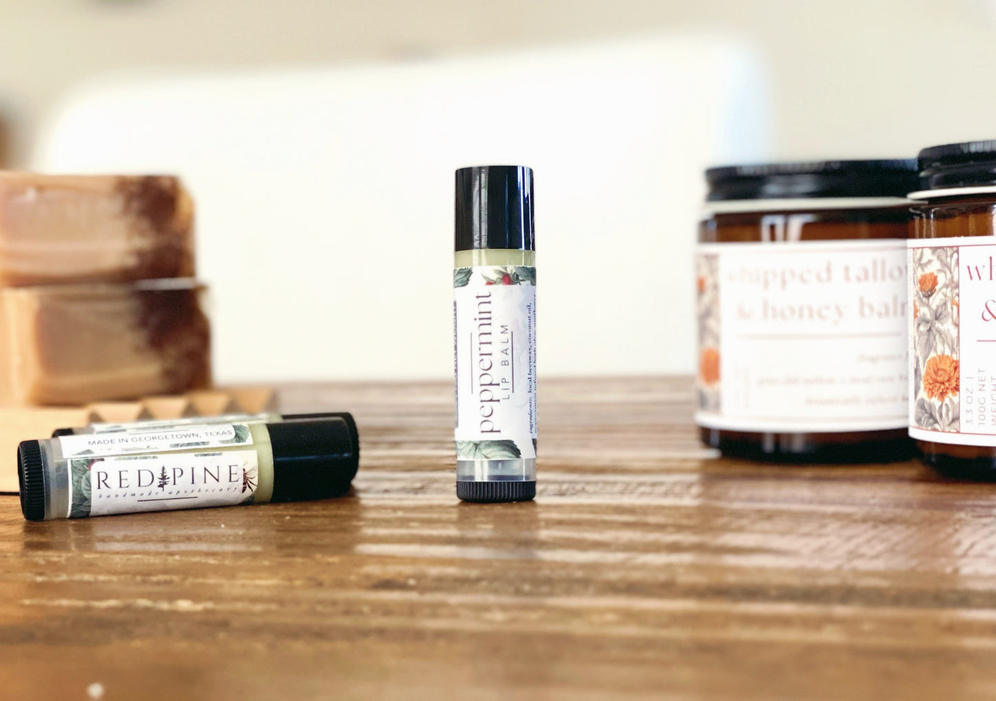 standing peppermint lip balm in the foreground on a wooden table with soap and whipped balm in the background.