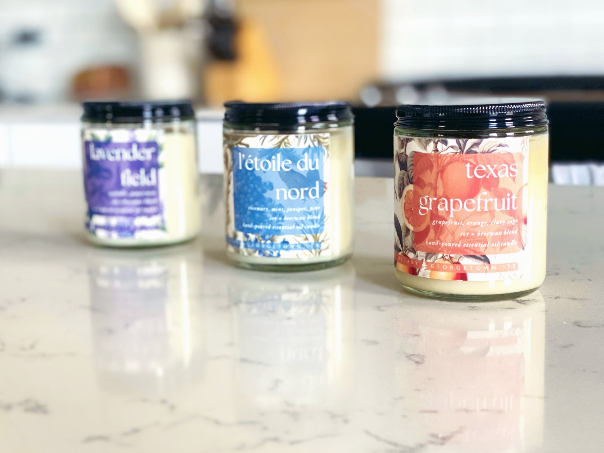 three candle jars are sitting on a quartz countertop in a white kitchen.  from left to right: lavender field, l'etoile du nord, texas grapefruit.