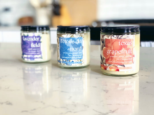 three candle jars are sitting on a quartz countertop in a white kitchen.  from left to right: lavender field, l'etoile du nord, texas grapefruit.