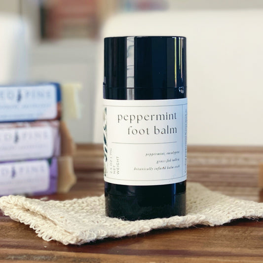 black tube of peppermint foot balm stick in the foreground on a wooden table.  there is a stack of soaps in the background.