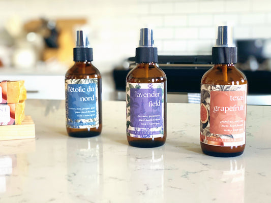 all three scents in the room spray collection are sitting on a quartz countertop in a white kitchen.  from left to right, l'etoile du nord, lavender field, texas grapefruit.