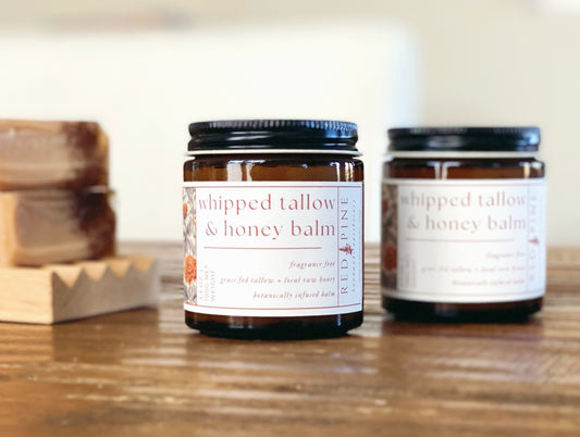 two jars of whipped tallow and honey balm in the foreground on a wooden table.  there are soap bars in the background.